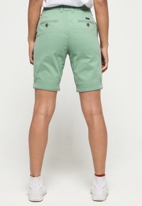 Muntgroene katoenen korte broek met opgerolde zoom, voorzien van achterzakken en twee knopen. Draagt wit sneakers.