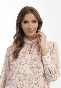 Lys pink bluse med blomstermønster, nøglehulshalsudskæring og rynkede ærmer. Stoffet har guld akcenter og en blød tekstur.