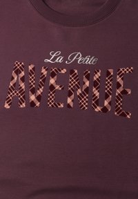 Sweat-shirt marron foncé avec "La Petite" en écriture argentée au-dessus de grandes lettres "AVENUE" en motif à carreaux rose et brun foncé sur le devant.