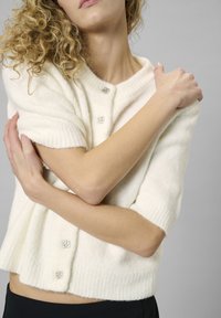 Valge kootud kardigan lühikeste varrukatega, millel on sooniktekstuur ja dekoratiivsed hõbedased nupud, mis on paigutatud esiosale.
