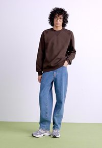 Brown sweatshirt with a loose fit, paired with light blue baggy jeans. Gray athletic shoes with white accents complete the outfit.