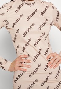 Beige jurk met lange mouwen en een aansluitend ontwerp, met een herhaald zwart Adidas-logo en trefoil-patroon, textuurstof met een lichte glans.