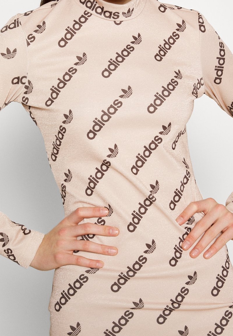 Beige jurk met lange mouwen en een aansluitend ontwerp, met een herhaald zwart Adidas-logo en trefoil-patroon, textuurstof met een lichte glans.