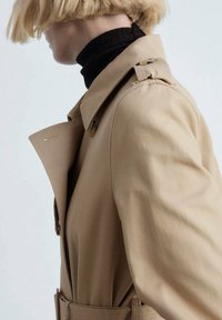 Trench coat beige avec un col classique, une ceinture latérale et des épaulettes discrètes. Porté sur un col roulé noir. Texture lisse.