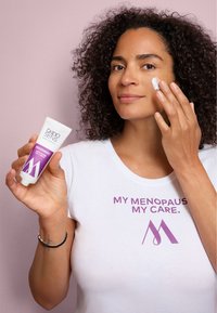 White tube of Dado Sens Menopausery Let’s Glow Dry Cream, with purple accents, held by a person applying cream to their face.