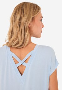 Top azzurro chiaro con maniche corte e un design a forma di V sul retro, caratterizzato da spalline incrociate. Realizzato in tessuto morbido con una consistenza liscia.