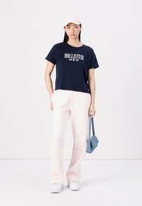 Námornícky crop tričko s textom "HOLLISTER", svetloružové široké nohavice, biele tenisky a svetloružová čiapka, drží malú modrú tašku.