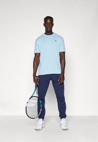 T-shirt di cotone azzurro chiaro con collo a giro, pantaloni della tuta blu navy e sneaker bianche. Tiene una racchetta da tennis con telaio nero e blu.