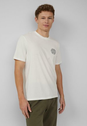 Biała bawełniana koszulka z krótkim rękawem, z okrągłym dekoltem i czarnym graficznym wzorem po lewej stronie klatki piersiowej. Noszona z oliwkowymi spodniami.