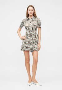 Γυναίκα που στέκεται με το χέρι στη μέση, φοράει φόρεμα τύπου wrap με κοντά μανίκια και γεωμετρικό σχέδιο, και λευκά παπούτσια με μυτερό μπροστινό μέρος και χαμηλό τακούνι, σε λευκό φόντο.