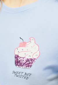 Μπλε πουκάμισο με γραφικό κέικ με ροζ παγιέτες, λευκή γλάσσο με στρας, μια λαμπερή κερασάκι και το κείμενο «Γλυκό αλλά στριφνό.»