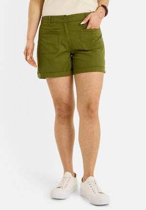 Mujer con pantalones cortos oliva con puños y bolsillos delanteros, zapatillas blancas y camiseta crema, de pie con una mano en el bolsillo.