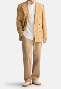 Beige dvouřadý blazer přes bílou košili, v kombinaci s béžovými kalhotami a krémovými teniskami. Hladká textura, strukturovaný design a klasický střih.