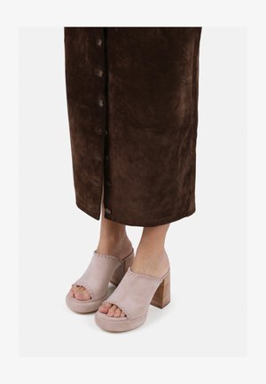 Halvány rózsaszín nubuk mules, nyitott orral és varrott részletekkel, fából készült tömbsarkon, egy barna nubuk gombos szoknyával viselve.