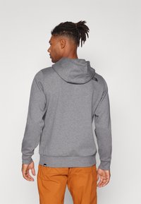 Grå sweatshirt med hætte, lavet af et blødt stof. Har et lille sort logo på ryggen og ribbede manchetter og kant. Sæt sammen med orange bukser.