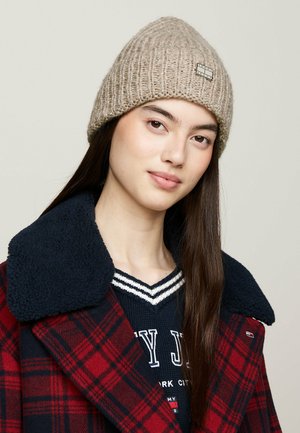 Gorro de punto beige con textura acanalada y un pequeño parche con logo. Llevado con un suéter azul marino y un abrigo de cuadros rojos con cuello de piel.