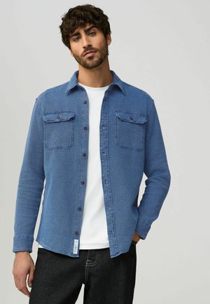 Chemise bleue à boutons, en tissu texturé, avec deux poches poitrine à rabat, boutons noirs, col, portée sur une chemise blanche, associée à un jean foncé.