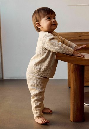 Dojenček stoji boso, se naslanja na leseno mizo, oblečen v bež teksturirano majico z dolgimi rokavi in ujemajoče hlače v notranjosti.