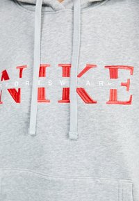 Pilkas džemperis su kapuce, pagamintas iš medvilnės ir sintetinių pluoštų, su raudonai siūtu "NIKE" tekstu ir baltu "SPORTSWEAR" tekstu krūtinėje. Įdėtas kapucės raištelis ir priekinė kišenė.