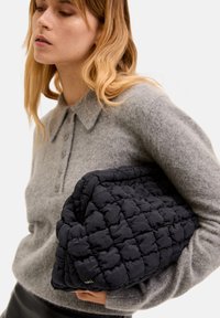 Mustat tilkkutyökangasta oleva laukku, jota pidetään kainalossa, yhdistettynä vaaleanharmaaseen villaneulepaitaan, jossa on kauluskanta.