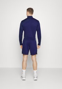 Compleu sportiv de culoare albastru închis, confecționat dintr-un material lucios, cu guler înalt, mâneci lungi și pantaloni scurți cu deschideri laterale. Adidași și șosete albe.