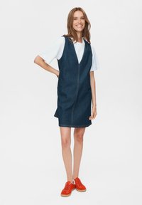 Denim mouwloze jurk met een V-hals, voorzien van een voorrits en zijsplitten. G gedragen over een wit T-shirt, gecombineerd met oranje sneakers.