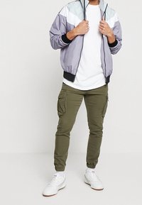 Ljuslila och grå bomberjacka, vit t-shirt, olivfärgade cargobyxor och vita sneakers. Jackan har elastiska ärmslut och dragkedja fram.