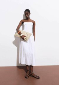 Witte strapless jurk van textuurstof, gecombineerd met een crèmekleurige clutch met franjes en zwarte platte sandalen met een decoratief accent.