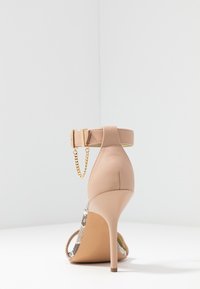 Nude högklackad sandal med en tunn ankelrem, guldkedjeaccent och en kombination av släta och texturerade material.