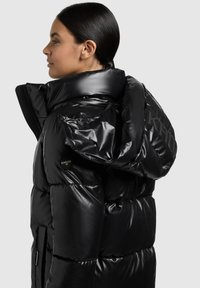 Crna jakna od puha s visokim ovratnikom i kapuljačom, sjajnog izgleda. Ima bočne džepove i logo na prsima i rukavu.