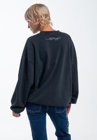 Felpa nera oversize con maniche raglan, tessuto strutturato, caratterizzata da un sottile logo ricamato sul retro. Indossata con jeans blu.