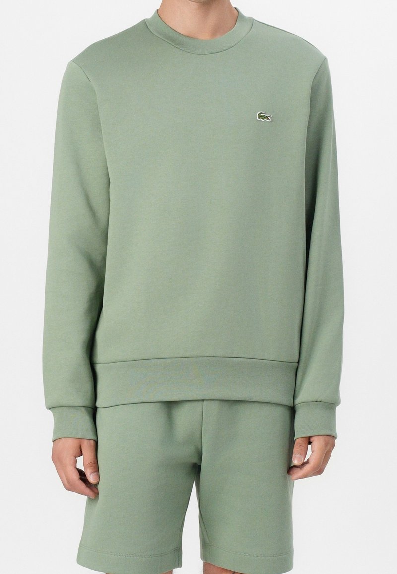 Homme portant un sweat-shirt vert clair à col rond et un short assorti avec un petit logo crocodile sur la poitrine.