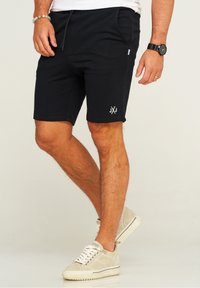 Black cotton shorts with an elastic waistband, side pockets, and white logo accent. Worn with beige sneakers, showing a sporty style.