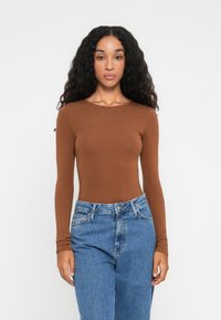 Kvinne med langt krøllete hår iført en tettsittende brun langermet topp og høytlivs blå jeans stående mot en ensfarget bakgrunn.