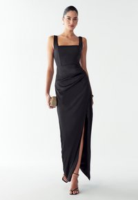 Žena v bezramínkových černých večerních šatech s rozparkem až k stehnům, držící béžovou kabelku a obutá v černých sandálech na tenkých řemíncích.