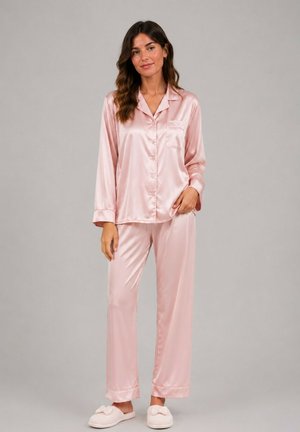 Femme debout portant un haut de pyjama en satin rose clair à manches longues boutonné et un pantalon assorti avec des chaussons blancs, fond gris neutre.
