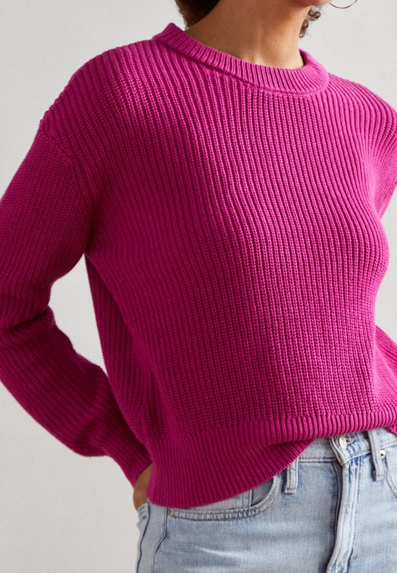 Femme portant un pull en maille côtelée magenta vif à manches longues et un jean taille haute bleu clair.