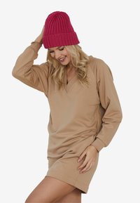 Beżowa sukienka dresowa z dekoltem w kształcie V i długimi rękawami. Noszona z jasnoróżową dzianinową czapką. Gładka tekstura, casualowy styl.