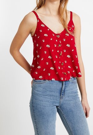Rotes ärmelloses Oberteil mit Blumenmuster und V-Ausschnitt, vorne mit Holzknöpfen und lockerem Schnitt. Kombiniert mit hellblauen Jeans.