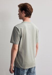 Hellgraues Baumwoll-T-Shirt mit kurzen Ärmeln und Rundhalsausschnitt, das eine lockere Passform und eine glatte Textur aufweist. Der Saum ist leicht abgerundet.