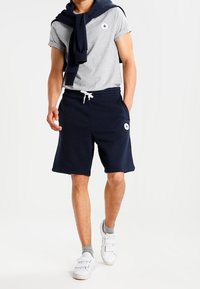 Camiseta gris con un suéter de cuello redondo azul marino atado alrededor de los hombros, combinado con pantalones cortos azul marino y zapatillas blancas.