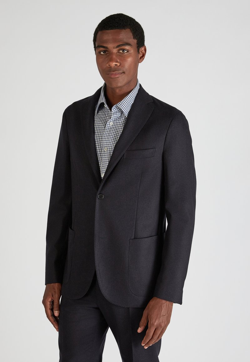 Ciemnoniebieska marynarka z dwoma przednimi kieszeniami i zapięciem na jeden guzik, noszona na jasnej koszuli w kratę. Gładka faktura i dopasowany krój.