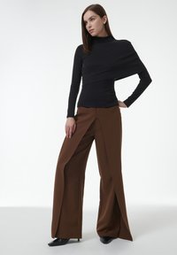 Schwarzes Langarmoberteil mit asymmetrischem Design und hohem Kragen; kombiniert mit braunen, weit geschnittenen Hosen mit einem vorderen Schlitz und glatter Textur.