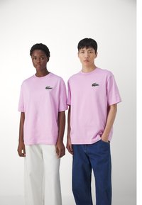 Due persone indossano t-shirt a maniche corte e dalla vestibilità comoda di colore rosa pastello, decorate con un logo ricamato verde. Lo sfondo è bianco uniforme.