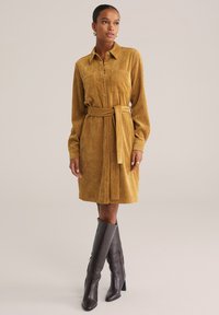 Rochie cămașă din corduroy muștar, cu talie accesorizată cu o curea, nasturi la față și două buzunare pe piept. Asortată cu bocanci din piele maro închis, până la genunchi.
