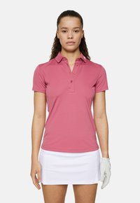 Roze poloshirt met een knoopsluiting bij de kraag, korte mouwen en subtiele textuur. Gecombineerd met een witte rok en een lichtgrijze golfhandschoen.