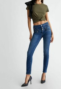Haut court noué vert olive avec manches à volants noires, associé à un jean skinny bleu foncé avec fermeture par boutons et coutures décoratives.