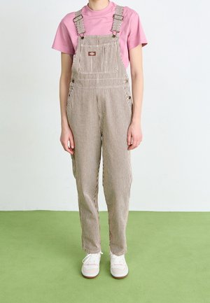 Salopette beige rayée avec bretelles, poche avant, surpiqûres contrastées, associée à un t-shirt rose et des baskets blanches sur un sol vert.