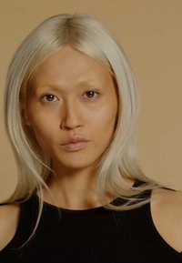 En person med langt, rett lyst blondehår iført et svart ribbestretch uten ermer, mot en lys beige bakgrunn.