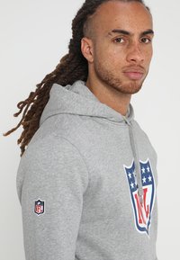 Sudadera gris hecha de tela suave, con un gran logo del escudo de la NFL en rojo, blanco y azul en la parte frontal y un parche de la NFL en la manga.